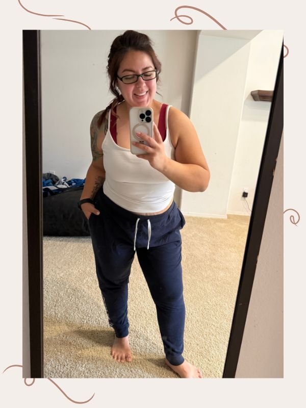 mirror selfie image of woman wearing dark blue joggers and white tank top with red bra straps showing