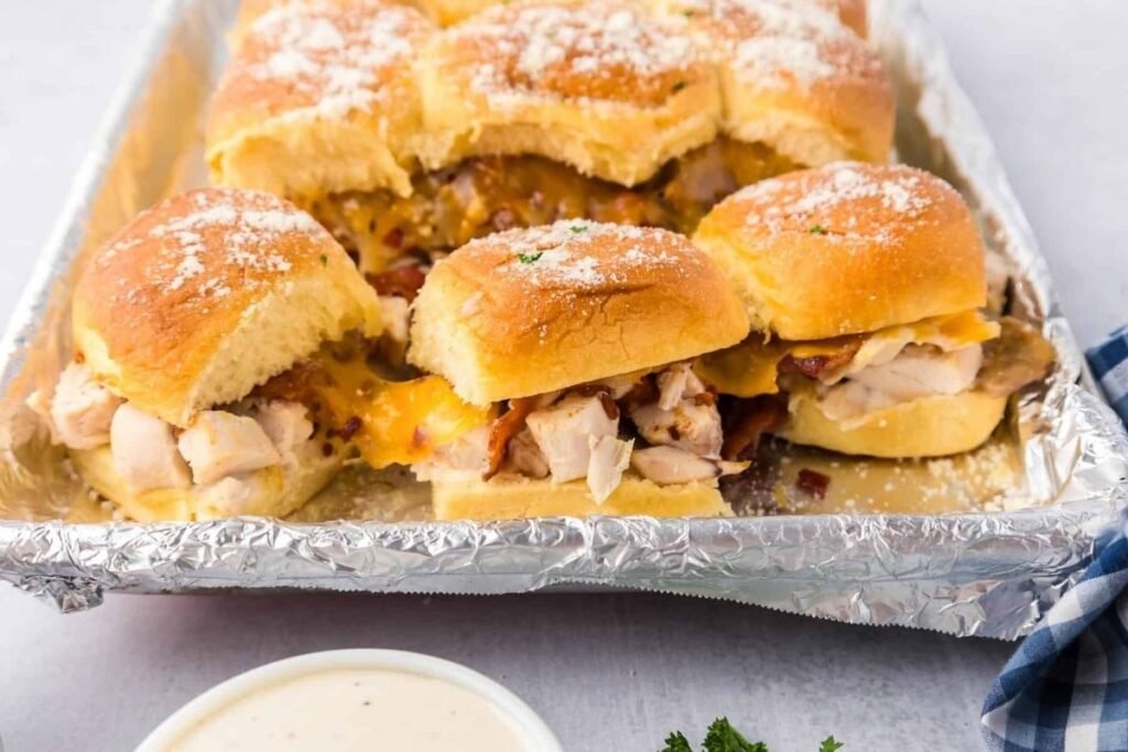 Close up image of baked chicken bacon ranch sliders on a sheet next to a bowl of ranch dipping sauce